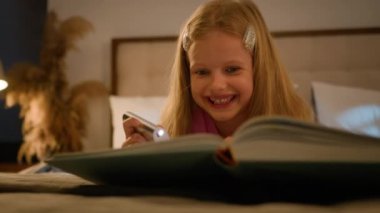 Smiling laughing cute little Caucasian girl preschooler child kid daughter reading book using flashlight in evening lying relaxing on bed enjoying read storybook funny stories fairytale story laugh