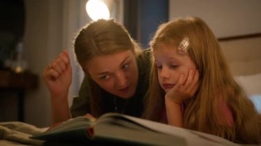 Happy Caucasian family loving caring mother reading book to little kid daughter using flashlight in bedroom lying on bed evening mom and child girl enjoying read fairytale story bedtime parenthood