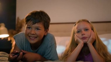 Smiling Caucasian children happy family kids boy girl brother sister siblings watching TV together watch cartoon show program using remote control enjoy television lying on bed in evening communicate