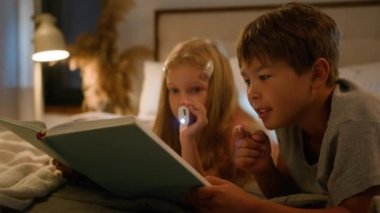 Caucasian children kids boy girl older brother younger sister lying on bed at home evening reading interesting book using flashlight read fairytale story for night bedtime happy family relationships