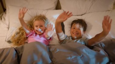 Funny playful laughing Caucasian children kids boy girl brother sister siblings happy family lying in bedroom on bed before bedtime waving hello greeting playing fooling around covered with blanket