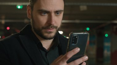 Otoparkta cep telefonu kullanan beyaz bir adam. İş adamı. İnternet zımbırtısı bağımlısı. Mesaj yazıyor.