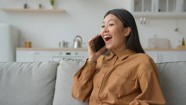 Excited Asian woman businesswoman talk phone in kitchen listen good news feel shock surprised korean chinese girl speaking cellphone enjoy mobile call discuss news laugh talking laughing win success