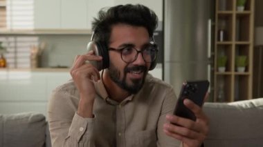 Tatmin olmuş mutlu, rahat Hintli Arap erkek ev sahibi kulaklıkla müzik dinliyor cep telefonu ses uygulamasında evde dinlenen online servis koltukta favori şarkıyı çalıyor.