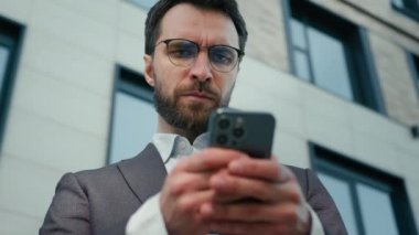 Ciddi bir Kafkasyalı girişimci yetişkin erkek işadamı CEO iş adamı ofis yöneticisi açık havada akıllı telefona bakarak şehirdeki sosyal ağları tarayıp cep telefonuyla sohbet ediyor.
