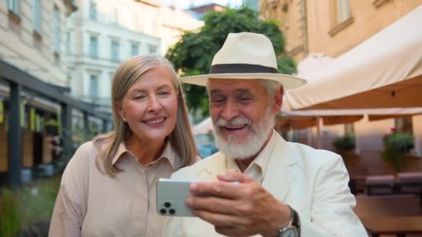 ハッピー笑顔のコーカサス高齢者カップルは 外の街の通りで一緒に話すガジェット携帯電話を笑って歩いています ソーシャルメディア ビデオ コール オンライン ブロッグ — ストック動画