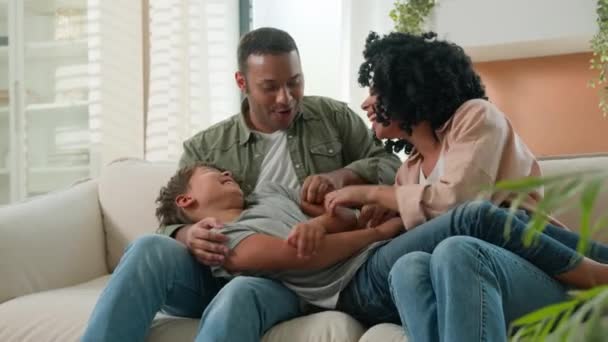 かわいい小さな男の子と遊ぶ幸せな多種多様な家族の両親は 楽しい笑いを一緒に家ソファの遊び心のあるアフリカ系アメリカ人のママのお父さんは子供の愛情関係を受け入れる — ストック動画