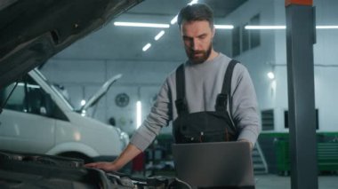 Araba servisi atölyesinde çalışan beyaz tenli tamirci, bilgisayar teknolojisiyle otomobil tamiri için elektronik dizüstü bilgisayarı kullanarak otomobil tamiri yapıyor.