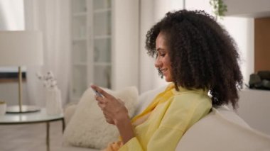 20 'li yaşlarda gülümseyen Afro-Amerikalı etnik kadın rahat ol akıllı telefonlar al internet siparişleri al mobil uygulamalar, telefon aleti bağımlılığı sosyal medya dairede