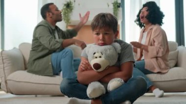 Üzgün küçük çocuk oyuncak ayıcığa sarılırken anne baba bağırarak kavga ediyor üzgün çocuk acı çekiyor anne baba boşanmada anlaşmazlık yaşıyor psikolojik travma velayeti çok ırklı ailede