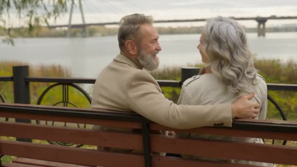 バックビュー幸せなシニア成熟した60S夫妻は ベンチ屋外で抱擁健康な退職カップル 中年女性の男は笑うリラックスしてロマンチックな週末を一緒に愛する都市公園自然の中で年齢 — ストック動画