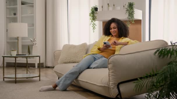 アフリカ系アメリカ人少女笑顔の若い女性は 家庭のスクロールスマートフォンでソファーで現代的なデバイスを使用して携帯電話を保持しています 笑うソーシャルネットワーク 女性のブロガーガジェット中毒オンライン購入 — ストック動画