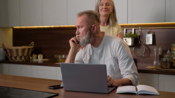 シニア 中年男性 ビジネスマン リモートワーキングコンピュータ キッチンでラップトップ思考の決定をオンラインで研究 笑顔の女性 サポート 自宅のコーカサス家族の愛 — ストック動画