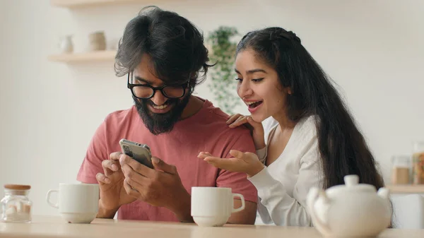 Excited multiethnic family Arabian indian man and woman looking at mobile phone screen at home kitchen happy couple girl and guy make order winning achievement shopping online with smartphone laughing
