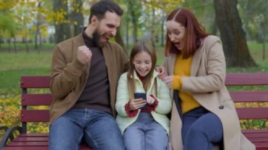 Mutlu Kafkas ailesi anne kızı, güz parkında bankta oturuyor cep telefonu kullanıyor ebeveynler ve küçük kız çocuğu başarıya ulaşıyor iyi haber akıllı telefon zafer tepkisi hayret verici