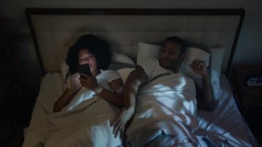 Sinirli Afro-Amerikalı erkek arkadaş uyumaya çalışıyor koca kızgın itme aleti bağımlısı eş internet bağımlısı kız arkadaş gece yatakta cep telefonlu sosyal medyada aile sorunları yaşıyor.