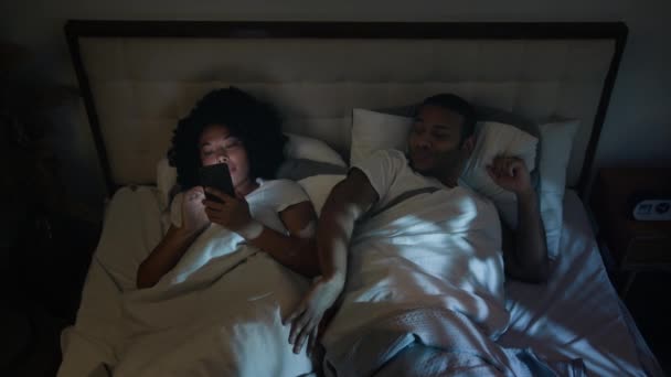 アフリカ系アメリカ人男性ボーイフレンド 睡眠をしようとしている夫怒っているプッシュガジェット中毒妻インターネット中毒ガールフレンド女性 ベッドカップル家族の問題で夜に携帯電話のソーシャルメディアをスクロール — ストック動画