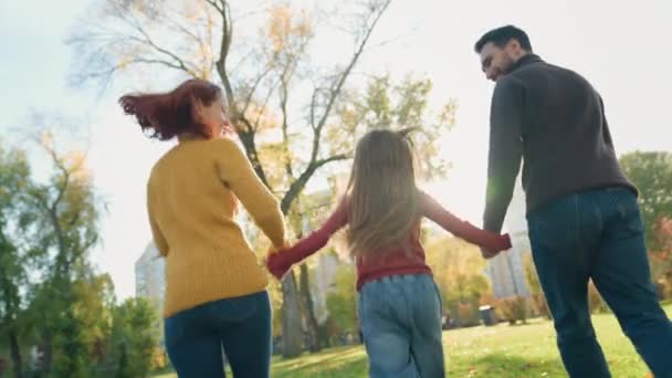幸せな家族の両親の母親の背後からの背景 草の秋公園の自然の中で手を握る娘の娘の子供の父親と一緒に休日を走る人々は楽しい親権の保育を再生します — ストック動画