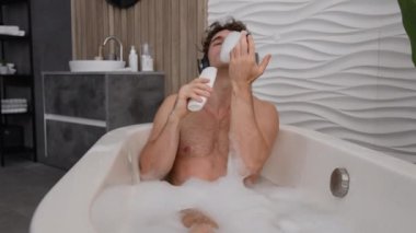 Kaygısız, komik, komik, beyaz, erkek, sıcak banyo suyunda tek başına dinleniyor. Evde dinleniyor, mikrofon gibi şarkı söylüyor, kulaklıkla müzik dinliyor.