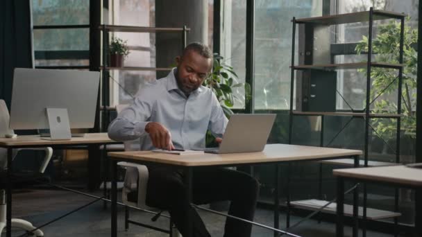 アフリカ系アメリカ人のビジネスマンマーケティングマネージャー 中年男性ライタージャーナリストがノートブックにテキストを書き込み ノートパソコンのチェックデータをオンラインで入力するビジネスプランデータ分析 — ストック動画