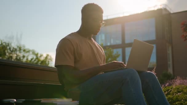 アフリカ系アメリカ人の男の学生は 大学のキャンパス近くの都市でコンピュータ男性の研究で屋外で勉強している男は 太陽の光の中でラップトップでオンラインで学ぶ宿題を考えて離れて離れて見ています — ストック動画