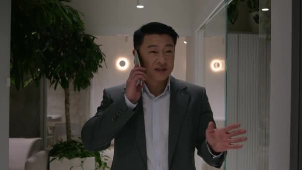 笑顔の中年アジアのビジネスマンがオフィスロビーを歩いています 男性プロ雇用主Ceo 韓国人ビジネスマン モバイル会話交渉スマートフォン通話 — ストック動画