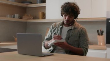 Kulaklıklı İspanyol adam, dizüstü bilgisayar konferansı, uzak sınıf koçluk dersi, e-öğrenme, Hint mutfağındaki Arap erkek işadamı, evde video konuşması yapan notlar yazıyor.
