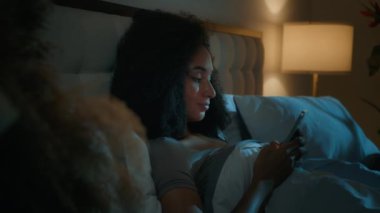 Güzel Afrikalı bir kadın, gece yatağında uzanmış akıllı telefon kullanıyor. Uykusuzluk çekiyor, cep telefonlarını dinlendiriyor, medya ağlarında geziyor, gülümsüyor, blog yazarı kız etkili alet bağımlısı, uyumadan önce.