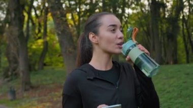 Fitness kadını, Kafkasyalı sporcu kız şehir parkında spor şişesiyle yürüyor, akıllı telefon kullanıyor, sosyal medyayı kontrol ediyor, sosyal medyada mutlu bayan blogcu sağlıklı dışarıda egzersiz yapıyor.