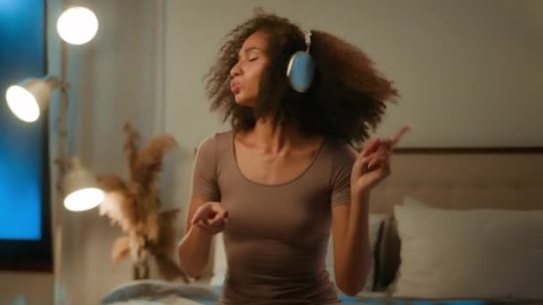 快乐的非洲裔美国女孩在床上听着音乐在耳机里播放音频音乐在黄昏灯下轻盈的少女在家里放松假装打鼓音乐节拍在耳机里享受音乐 — 图库视频影像