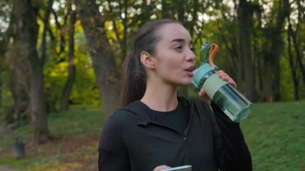 健康女性白种人运动女孩带着运动瓶装水在城市公园里散步 使用智能手机检查手机社交媒体快乐女性年轻博主在室外训练健康运动 — 图库视频影像