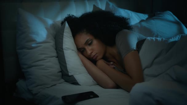 自宅でベッドで寝ている美しいアフリカ系アメリカ人の少女が目を覚ますと 警報時計の携帯電話のメッセージは 着信するスマートフォン通話で邪魔された夜に怒っている民族の女性を怒らせました — ストック動画