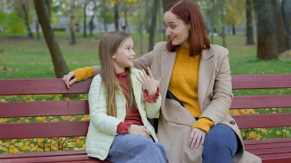 Caucasian woman mother mom talking with child girl kid daughter teen schoolgirl family talk friendly conversation motherhood childhood childcare sitting at city park autumn at bench laugh smile news
