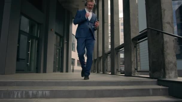 アクティブな面白い移動ダンス古いビジネスマン幸せな中年コーカサス成熟したビジネスマンは 街の階段オフィスを歩いているイヤホンと電話でヘッドフォンで音楽を聞く歌にダンスを歩きます — ストック動画