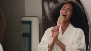 Banyodaki kıvırcık saçlı mutlu Afrikalı kadın aynaya bakıyor nemlendirilmiş cilt bakımının tadını çıkarıyor saç bakımını şımartıyor banyo fırçasında mikrofon hijyenik rutin gibi şarkı söyleyerek şımartıyor