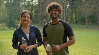 Doğa parkında açık hava portresi Hintli Arap çift arkadaş, kadın kız, etnik evli sporcu sporcular spor yaptıktan sonra elinde su şişeleriyle kameraya gülümseyen sporcular.