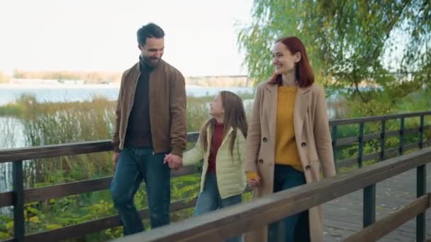 快乐的白种人父母夫妇收养了可爱的女儿女儿女儿周末在公园秋天户外湖边小桥边散步边谈笑风生手牵手散步 — 图库视频影像