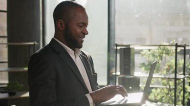 Üst düzey Afro-Amerikan etnik işadamı güneş ışınlarında dizüstü bilgisayarla şirket ofisinde duruyor. Gülümseyen iş adamı yöneticisi dijital veri tarama bilgisayar siteleriyle çevrimiçi çalışıyor