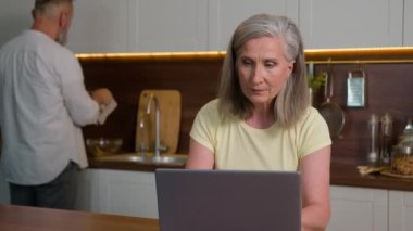 Orta yaşlı bir kadın, yetişkin bir iş kadını internette bilgisayarla çalışıyor. İş yerinde çalışan kadın dizüstü bilgisayar öğreniyor. Bu arada kıdemli adam mutfakta bulaşık yıkıyor.