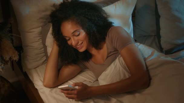 快乐的非洲裔美国女性用户电子产品成瘾社交媒体滚动手机在晚上的卧室睡觉微笑20多岁的女性失眠放松在床上浏览智能手机应用程序 — 图库视频影像