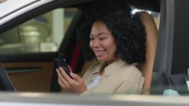 Afro-Amerikalı kadın şoför gülümseyen iş kadını akıllı telefon kullanarak park yerinde oturuyor ve cep telefonu ekranına bakıp yeni bir araba seçiyor.
