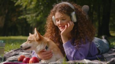 Mutlu portre evcil hayvanı ve sahibi gülümseyen beyaz kız kulaklıkla müzik dinliyor güzel bir köpek ile battaniyeye uzanmış Corgi açık hava genç kadın haftasonu şehir parkında yavru köpeğe sarılıyor.