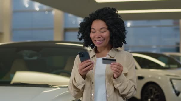 アフリカ系アメリカ人女性の顧客使用携帯電話自動車ショールーム車サロンディーラーストアで新しい高級車を購入し 自動車の簡単なローンクレジットハッピークライアントのお祝いのための銀行カードを支払う — ストック動画