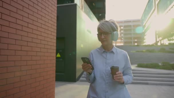 コーカサス 成熟した シニア 幸せな ビジネスマン ヘッドフォンで音楽を聞く 屋外で日当たりの良い日 携帯電話のスマートフォンを使用して コーヒー休憩 市内のビジネス — ストック動画