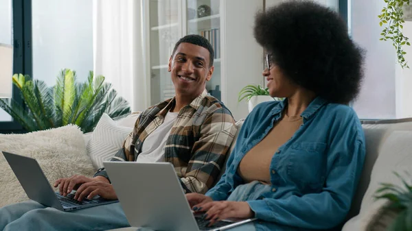 African American woman work with laptop man computer working couple sit at row two freelancers with computers coworking at home addict family looking at each other laughing playing game online on sofa
