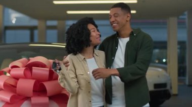 Afrikalı Amerikalı kadın erkek mutlu bir milenyum çifti müşterileri kameraya yeni arabanın anahtarını gösteriyor modern galeride kırmızı kurdeleli lüks otomobil galeride araba satın alıyor.