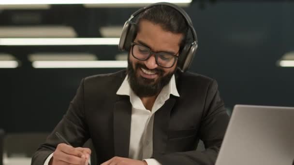 ハッピー笑顔満足のアラビアの男ビジネスマネージャー起業家雇用者インドの肯定的な笑顔のビジネスマンはノートブックでノートを書くヘッドフォンを身に着けます オフィスで勉強するオンラインレッスンクラスのビデオ通話 — ストック動画