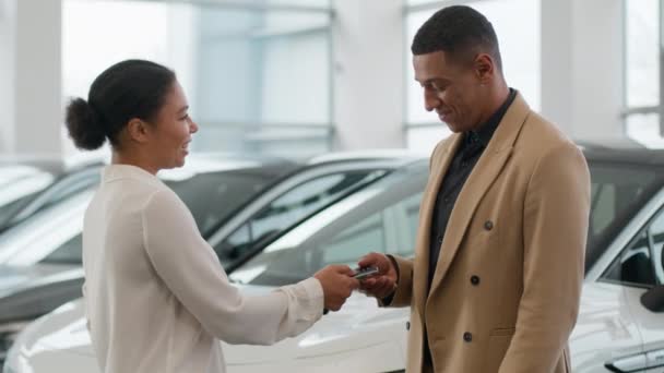 ハッピークライアントアフリカ系アメリカ人男性購入新しい車の笑顔女性販売代理店は ショールームレンタカー輸送成功販売で販売ディーラーのサロン自動車の高級車にキーを与えます — ストック動画