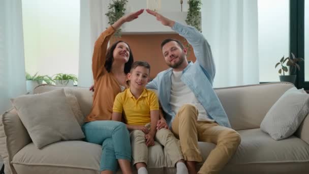 快乐的白种人家庭房主母亲父亲儿子父母小孩坐在舒适的客厅沙发上 手拿着房子的屋顶符号看着相机新租的房屋贷款 — 图库视频影像