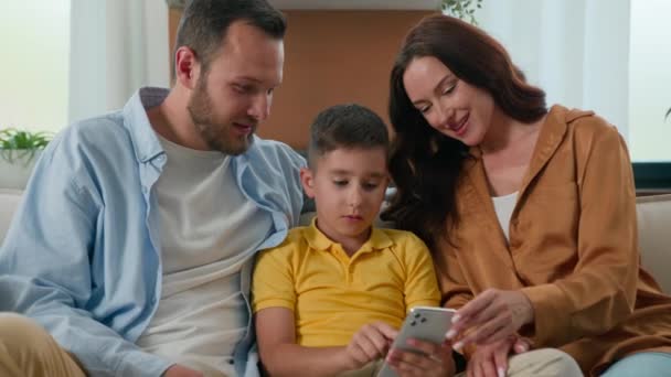 幸せな家族の父親の両親の息子の男の子の子供の話は 携帯電話でソーシャルメディアを閲覧するインターネット上でビデオを再生するためのモバイルアプリを使用して ソーシャルホームで一緒にスマートフォン画面を見ながらコミュニケーションします — ストック動画